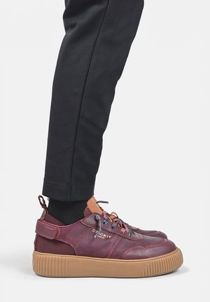 Persoon die zwarte broek en bordeauxrode sneakers draagt met tan dikke rubberen zolen en meerkleurige veters, staand op een witte achtergrond.