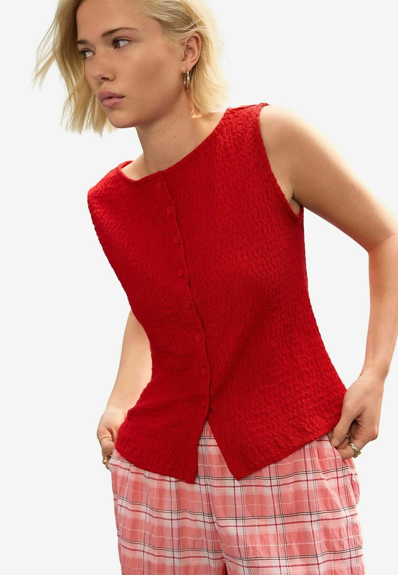 Blouse sans manches rouge en tissu texturé avec un col arrondi, dotée d'une rangée centrale de boutons et d'un ourlet évasé.
