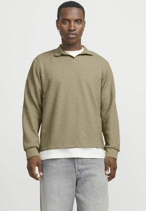 Khaki long-sleeve polo shirt with ribbed texture, a collared neck, and cuffs, layered over a white shirt, paired with light gray jeans.