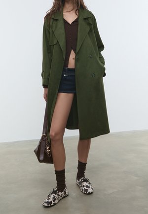 Une femme porte un long trench-coat vert olive par-dessus un haut marron court et un short en jean foncé, tenant un sac marron avec des chaussures à motif et des chaussettes foncées.