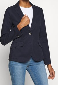 Blazer bleu marine en coton avec une coupe ajustée, doté d'une fermeture à un bouton, de deux poches avant et d'une texture lisse.