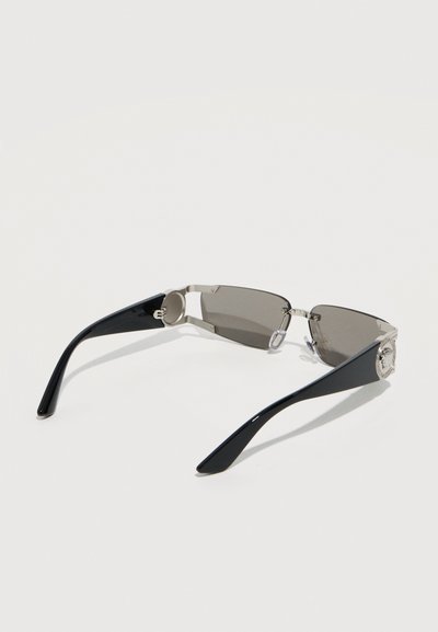 Κομψά γυαλιά ηλίου χωρίς πλαίσιο με σκουρόχρωμα φακούς και μαύρα καμπυλωτά μπράτσα που φέρουν ασημένια κυκλικά λογότυπα κοντά στις μεντεσέδες, τοποθετημένα πάνω σε απλό φόντο.