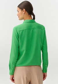 Camicia verde a maniche lunghe con colletto, texture liscia e polsini abbottonati. Presenta una vestibilità dritta e un dettaglio sulla parte posteriore.