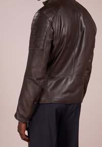 Brun läderjacka med en åtsittande design, med kviltade axeldetaljer och sidogettyrar. Slät textur, strukturerad silhuett.