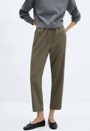 Person iført olivengrønne snor-bukser, grå kort sweater og sorte ballerinaer, stående med hænderne på hofterne.