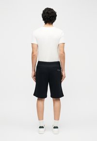Hombre con cabello rizado de pie de espaldas, vestido con camiseta blanca, pantalones cortos negros hasta la rodilla, calcetines blancos y zapatillas blancas.