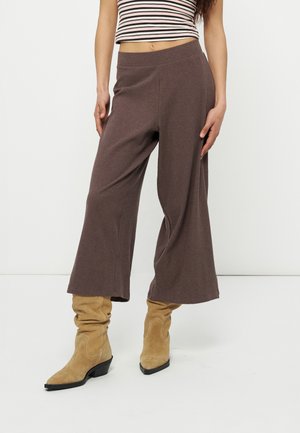 Kvinde iført brune, vidde, cropped bukser, beige slappe ruskindsstøvler og en stribet, ærmeløs croptop, der står mod en hvid baggrund.