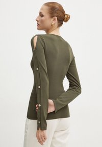 Maglione verde oliva a maniche lunghe con aperture sulle spalle, dotato di bottoni lungo le maniche e un design aderente. Tessuto morbido e elastico.