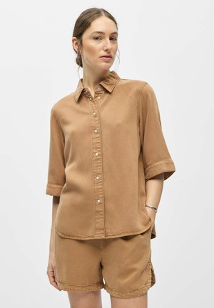 Femme portant une chemise boutonnée couleur beige à manches trois-quarts et un short assorti, debout avec une main dans la poche sur un fond blanc.