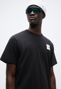 Μαύρο βαμβακερό μπλουζάκι με κοντά μανίκια, με λευκή σήμανση λογότυπου στην αριστερή πλευρά του στήθους. Φοριέται με γκρι σκουφάκι και αντανακλαστικά γυαλιά ηλίου.