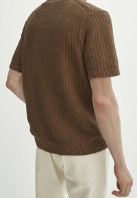 Brun strikket kortærmet sweater med et struktureret mønster, ribbet krave og kant, vist fra bagsiden mod en lys baggrund.