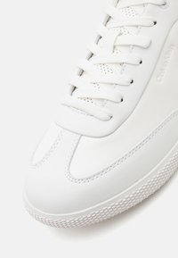 Detailní záběr na bílé tenisky Calvin Klein s texturovanou podrážkou, kulatou špičkou a bílými šňůrkami na hladkých a perforovaných horních panelech.