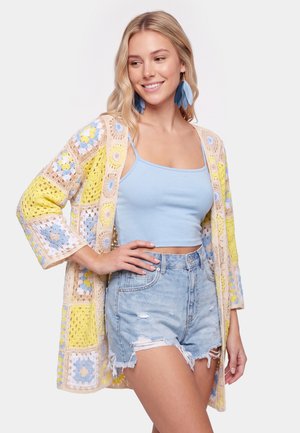 Lyshåret kvinde smilende, iført store blå øreringe formet som blade, en lyseblå crop top, denimshorts og en flerfarvet hækletrøje.