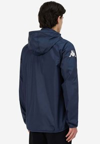 Uomo che indossa un giubbotto con cappuccio blu scuro, rivolto di spalle, con un logo bianco sulla manica sinistra superiore e pantaloni neri.