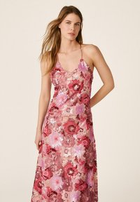Femme portant une robe rose à fleurs sans manches avec fines bretelles, debout devant un fond beige uni, une main sur la hanche.