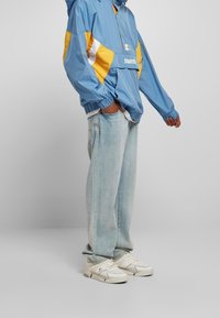 Giacca leggera blu con accenti gialli e bianchi, jeans leggeri azzurri a vestibilità rilassata e sneakers bianche con dettagli argentati.
