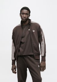 Καφέ αθλητική φόρμα με ζακέτα φερμουάρ και λευκές ρίγες, παντελόνι ταιριαστό, και μικρό λογότυπο στο στήθος και στο μηρό.