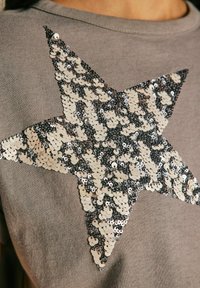 Chemise grise à manches courtes avec un grand motif d'étoile en sequins argent et noir alternés. Texture de tissu douce.
