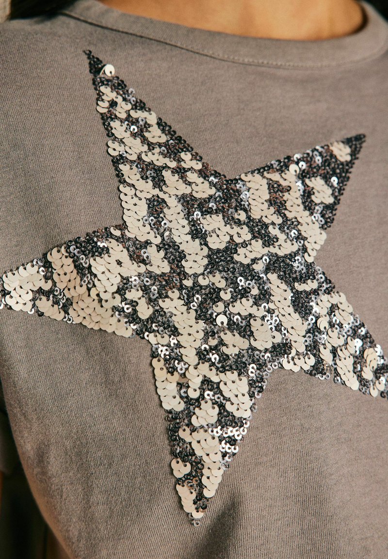 Chemise grise à manches courtes avec un grand motif d'étoile en sequins argent et noir alternés. Texture de tissu douce.
