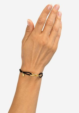 Braccialetto in corda nera con un anello di catena dorata e un piccolo ciondolo dorato, indossato su una mano con smalto neutro.