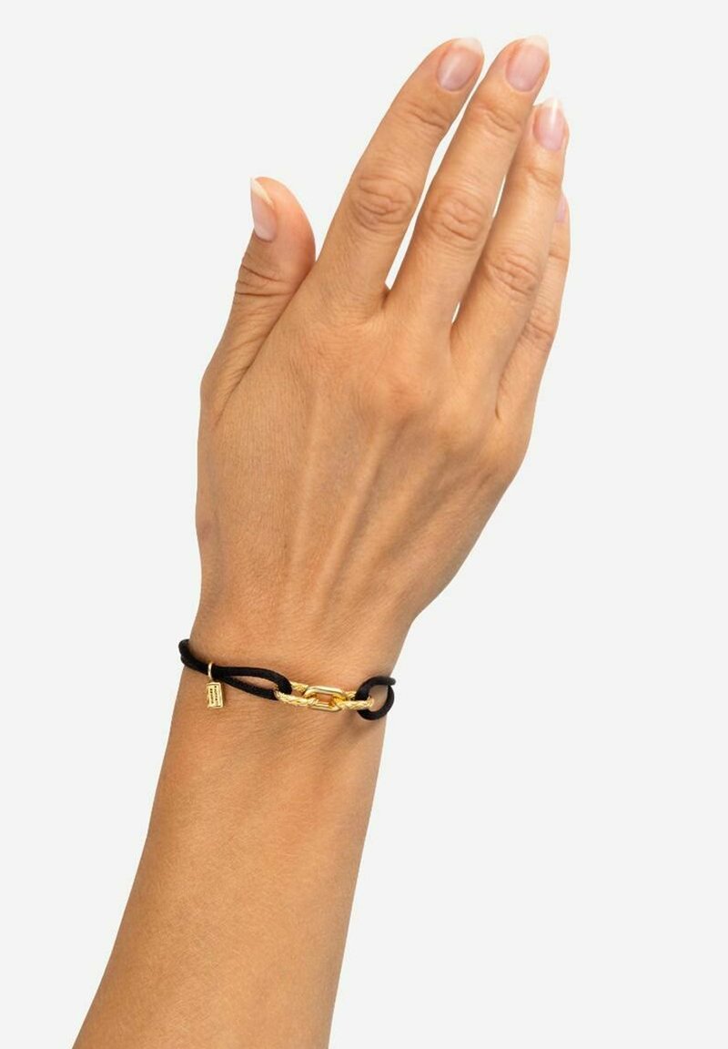 Braccialetto in corda nera con un anello di catena dorata e un piccolo ciondolo dorato, indossato su una mano con smalto neutro.