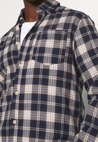 Granatowa koszula w kratę z beżowymi i białymi akcentami, z kieszonką na piersi, kołnierzykiem zapinanym na guziki oraz lekkim, teksturowanym materiałem.