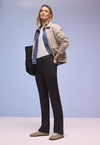 Joven mujer con chaqueta beige sobre camisa vaquera azul y camiseta blanca, pantalones negros y zapatillas beige, de pie con una mano en el bolsillo.