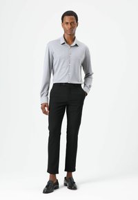 Grijs shirt met lange mouwen en knoopdetails, gecombineerd met nauwsluitende zwarte broek en zwarte schoenen, staand tegen een effen witte achtergrond.