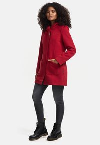 Manteau à capuche en mélange de laine rouge avec fermeture éclair sur le devant, deux poches plaquées et fentes latérales, associé à un jean skinny foncé et des bottes à lacets noires.