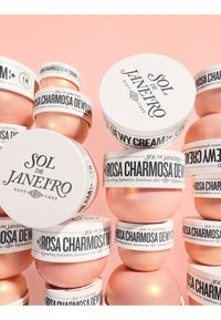 Behälter der Sol de Janeiro Rosa Charmosa Dewy Cream, mit weißen Etiketten, schwarzem Text und pinken Metallic-Akzenten, gestapelt vor einem pinken Hintergrund.