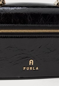 Melna struktūrēta ādas rokassoma ar zeltītu rāvējslēdzēju un piederumiem, ar izteiksmīgu zelta "FURLA" logo priekšā.
