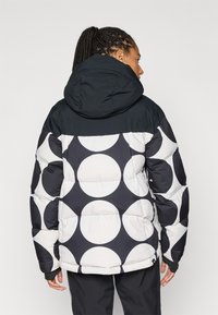 Pufferjack in zwart-wit met grote cirkelvormige patronen, voorzien van een capuchon en een contrasterend bovenpaneel. Getextureerd, geïsoleerd ontwerp.