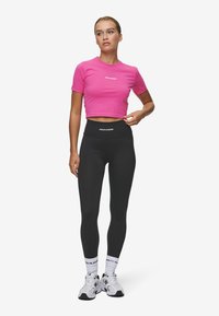 Ružičasti krojeni crop top s kratkim rukavima na kojem se nalazi logo "PEGADOR", u kombinaciji s crnim tajicama visokog struka i bijelim tenisicama.