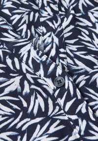Camicia blu navy con un motivo a foglia bianca, dotata di chiusura con bottoni e una texture di tessuto liscia. Cuciture dettagliate visibili.