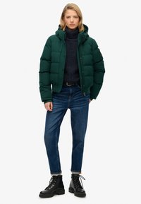 Zielona kurtka puchowa z kapturem, zapinana na zamek i zatrzaski, noszona na ciemnym swetrze. Dopełnieniem stylizacji są niebieskie dżinsy oraz czarne wiązane buty typu combat.