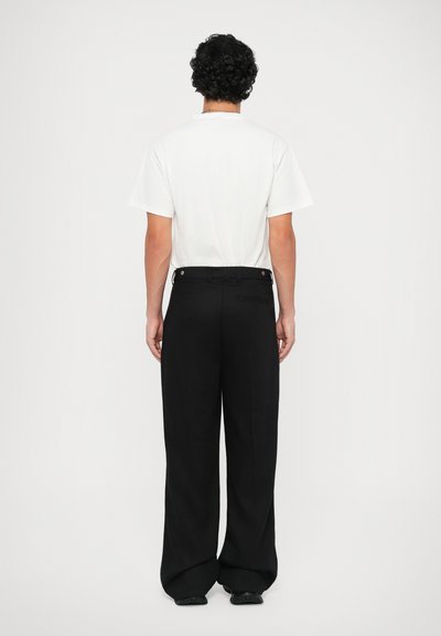 Weißes T-Shirt, schwarze weit geschnittene Hose, strukturierter Stoff, sichtbare Nähte und Verschlüsse an der Taille, von hinten betrachtet.