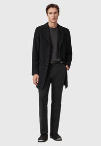 Mann mit Händen in den Taschen, der einen schwarzen gemusterten Mantel, einen dunkelgrauen Pullover, schwarze Hosen und schwarze Schuhe vor einem hellgrauen Hintergrund trägt.