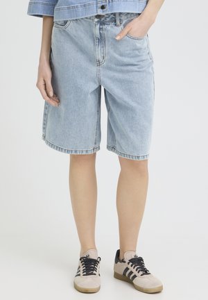 Személy világoskék farmer hosszú rövidnadrágot, világoskék farmer dzsekit és bézs színű, fekete csíkos tornacipőt visel, miközben egyszerű háttér előtt áll.