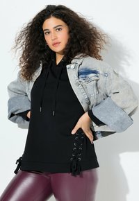 Donna con capelli ricci che indossa un giubbotto di denim chiaro sopra un hoodie nero con lacci laterali e leggings bordeaux lucidi, posando contro un muro bianco.