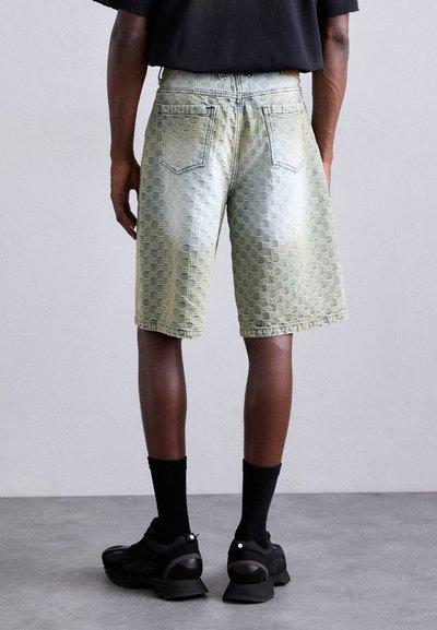 Homme portant un short en denim vert clair à motifs, des chaussettes noires et des baskets noires, se tenant sur un sol gris contre un mur blanc.