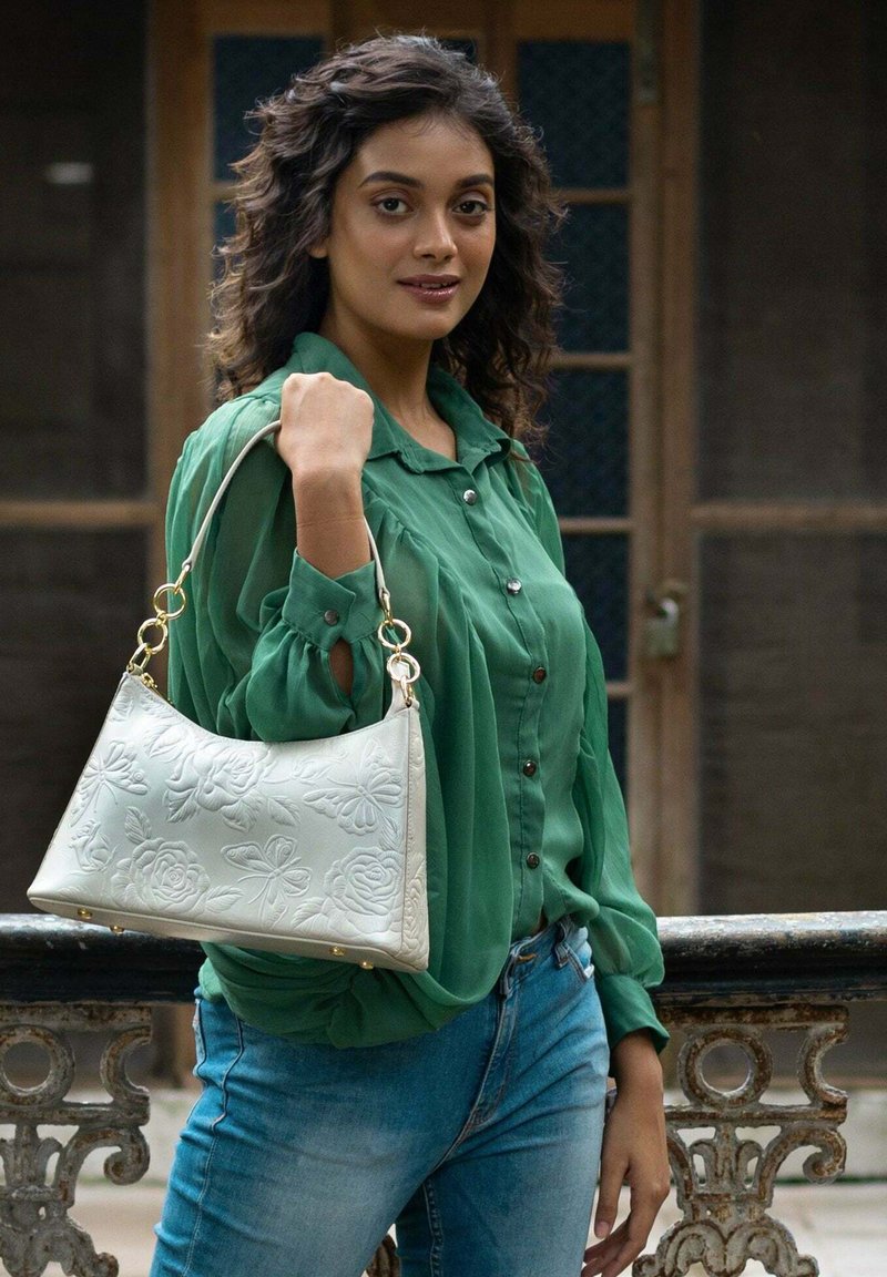 Weiße Lederhandtasche mit geprägtem Blumen- und Schmetterlingsmuster, goldenen Beschlägen und einem einzelnen Schultergurt, gehalten von einer Person in einer grünen Bluse.