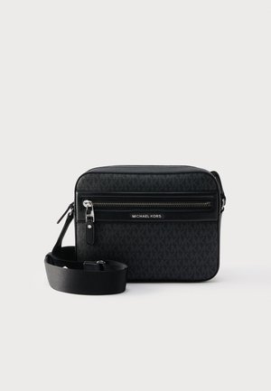 Sac bandoulière compact noir avec sangle réglable, poche avant zippée et logo Michael Kors embossé sur une surface à motifs.