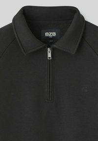 Pull noir avec col zippé, étiquette de marque BZB visible à l'intérieur, et un logo en lettres "B" en relief subtil sur la poitrine.