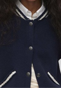 Marineblauer Cardigan mit weißen Streifenakzenten, rundem Kragen und Metallknöpfen. Verfügt über ein strukturiertes Strickgewebe sowie seitliche Taschen mit dekorativen Kanten.