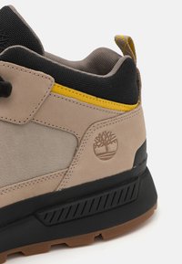 Beige velúr és textil sneaker sárga kiegész�ítőkkel, fekete hálós nyakkal és gumitalppal, amely textúrált mintázattal és logó részletezéssel rendelkezik.