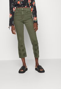Jeans cropped verde caqui com bordado floral na bainha. Usados com sandálias pretas abertas. O tecido é denim; o design é justo com uma cintura alta.