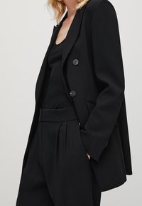 Femme portant un blazer noir ajusté à deux boutons, un top noir sans manches et un pantalon noir plissé taille haute, main dans la poche.