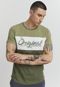 Zelena i bijela majica kratkih rukava s okruglim izrezom. Sadrži natpis "Original Jeansmaker" u kontrastnom fontu. Glatka tkanina.