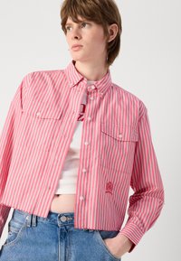Rosa och vit randig kortärmad skjorta med knäppning, två bröstfickor och broderade detaljer. Bärs över en vit topp och blå jeans.