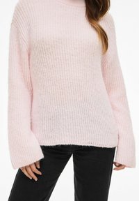 Różowy, duży sweter z dzianiny o teksturowanym wzorze żeberkowym, z szerokimi rękawami i wysokim kołnierzem. Zestawiony z czarnymi spodniami.